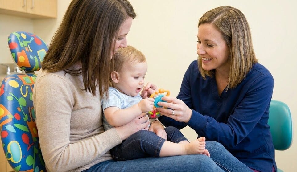 A friendly pediatric dentist gently examining a one-year-old child on their mother's lap during a first dental visit.