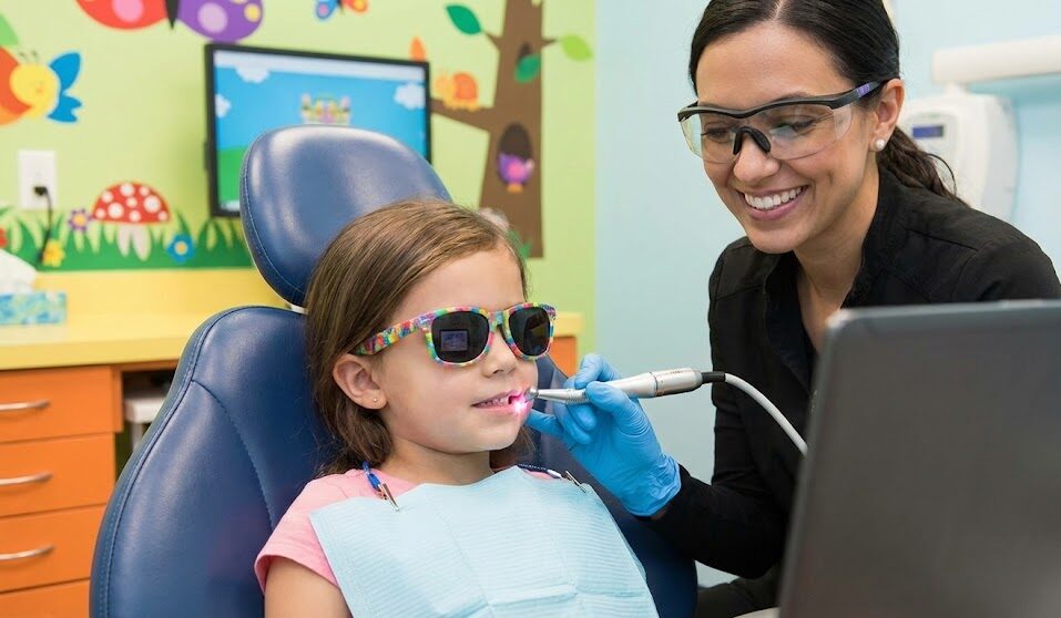 A smiling pediatric dentist using a dental laser on a young child in a colorful, child-friendly dental office.
