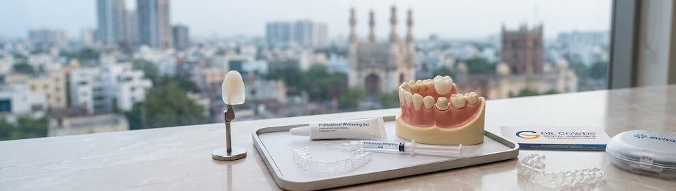 An arrangement of key cosmetic dentistry treatment options, featuring a single translucent porcelain veneer, a custom take-home whitening tray with gel, a Zirconia crown on a molar model, and a discreet clear aligner, all placed within a modern dental clinical environment at Dr. Gowd Dental Hospitals.